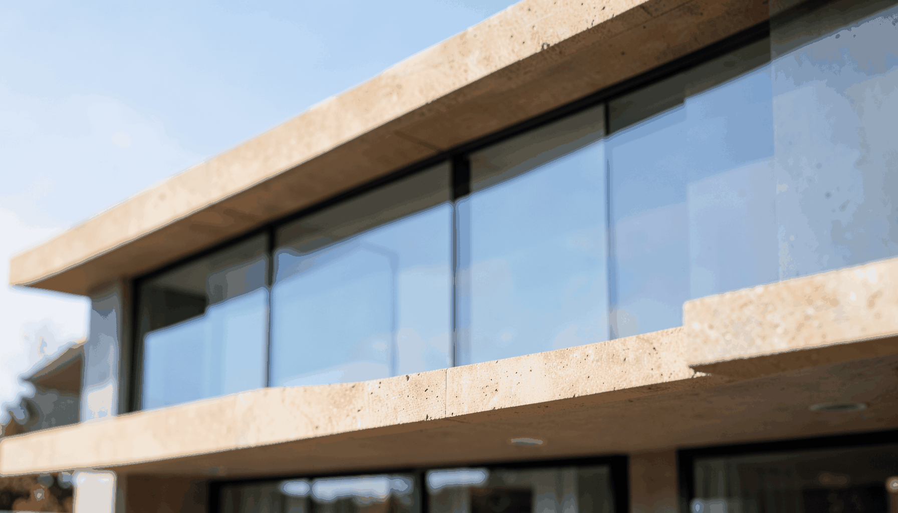 A crisp architectural photograph of a modernist house facade with clean horizontal lines, warm concrete tones, and glass panels reflecting a pale sky
