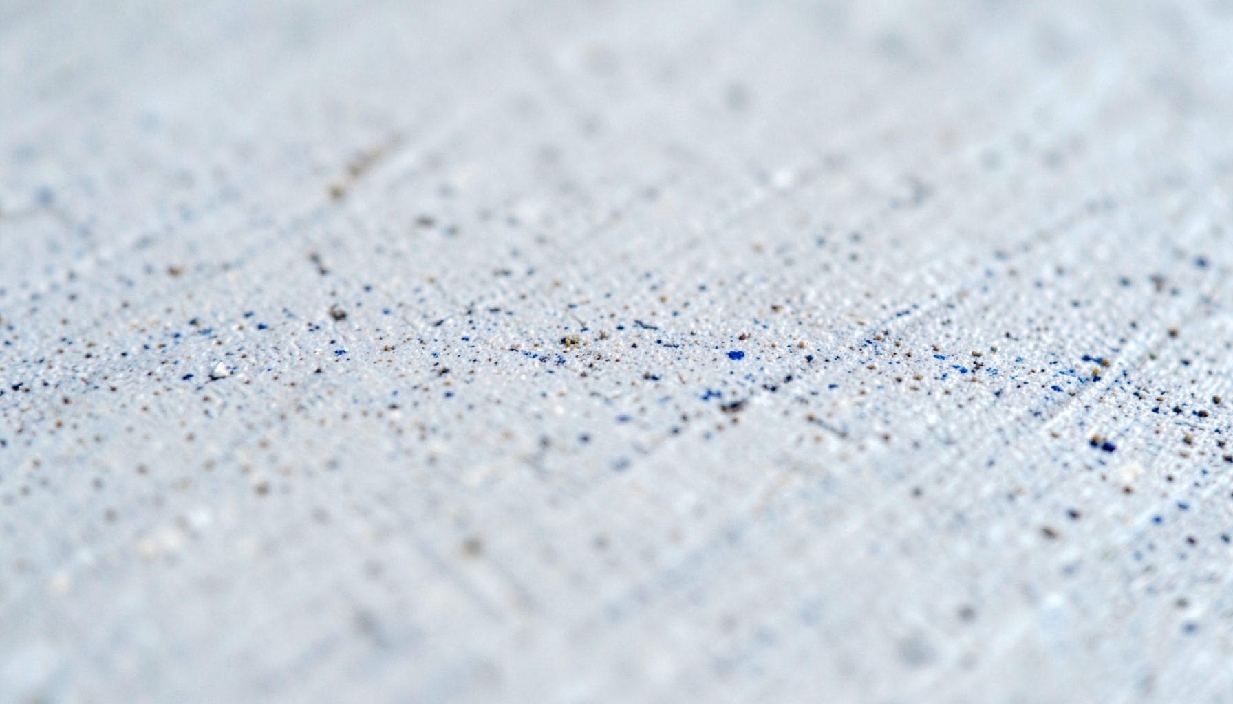 A tight macro shot of brushed concrete surface, pale grey with fine sandy speckles and subtle shadow lines.
