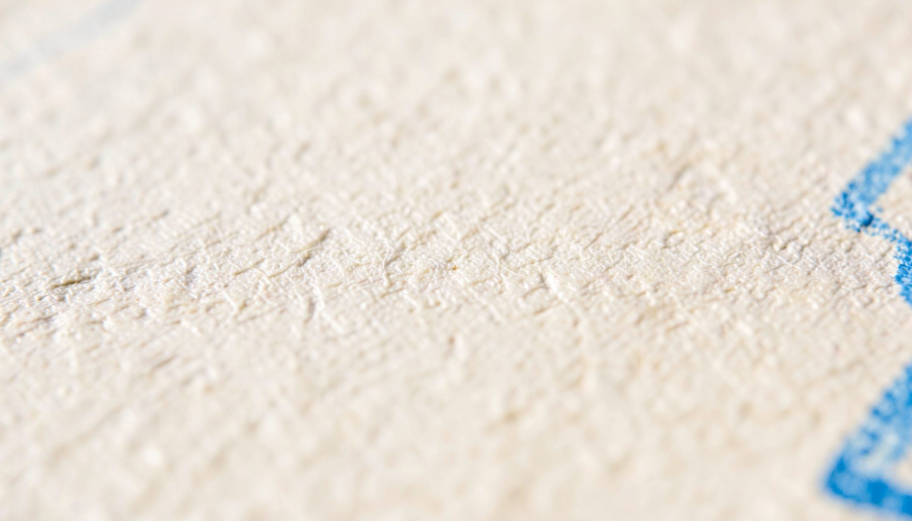 A tightly cropped macro photograph of matte paper grain, showing subtle fiber texture in warm off-white tones with faint shadows pooling in the crevices.