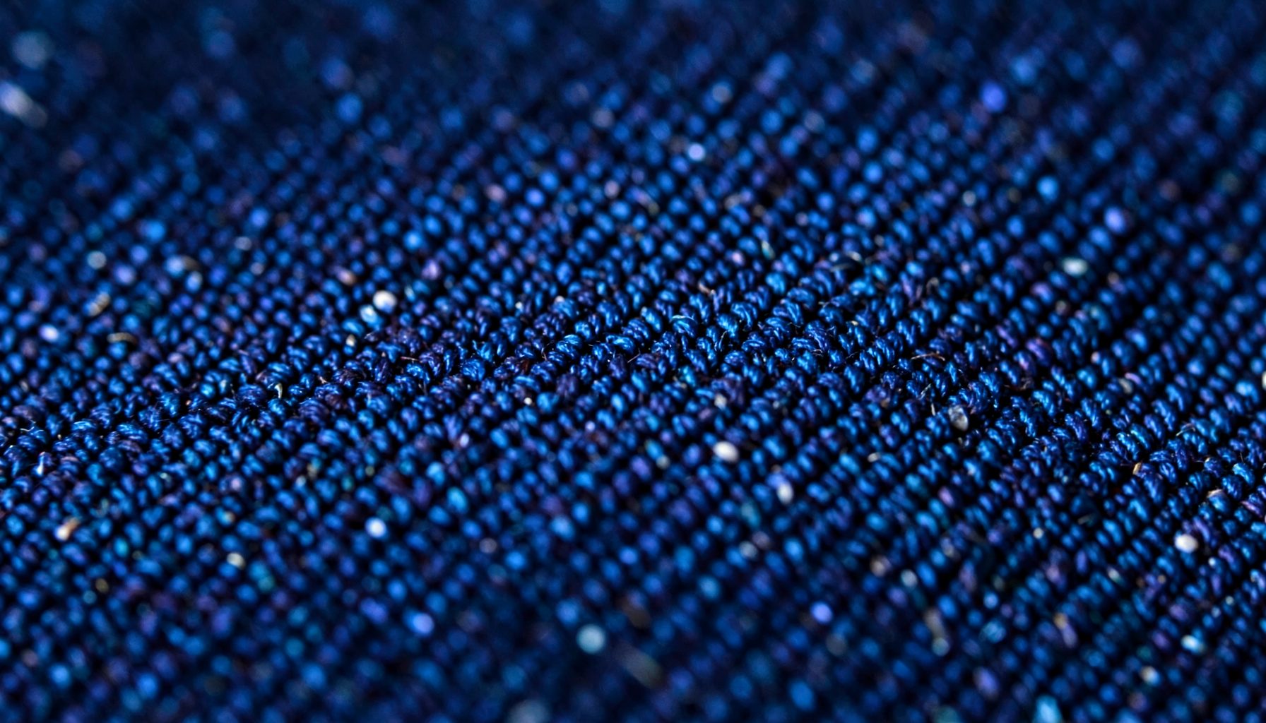 An extreme close-up of dark denim twill, deep indigo blue with diagonal weave ridges and faint white flecks.
